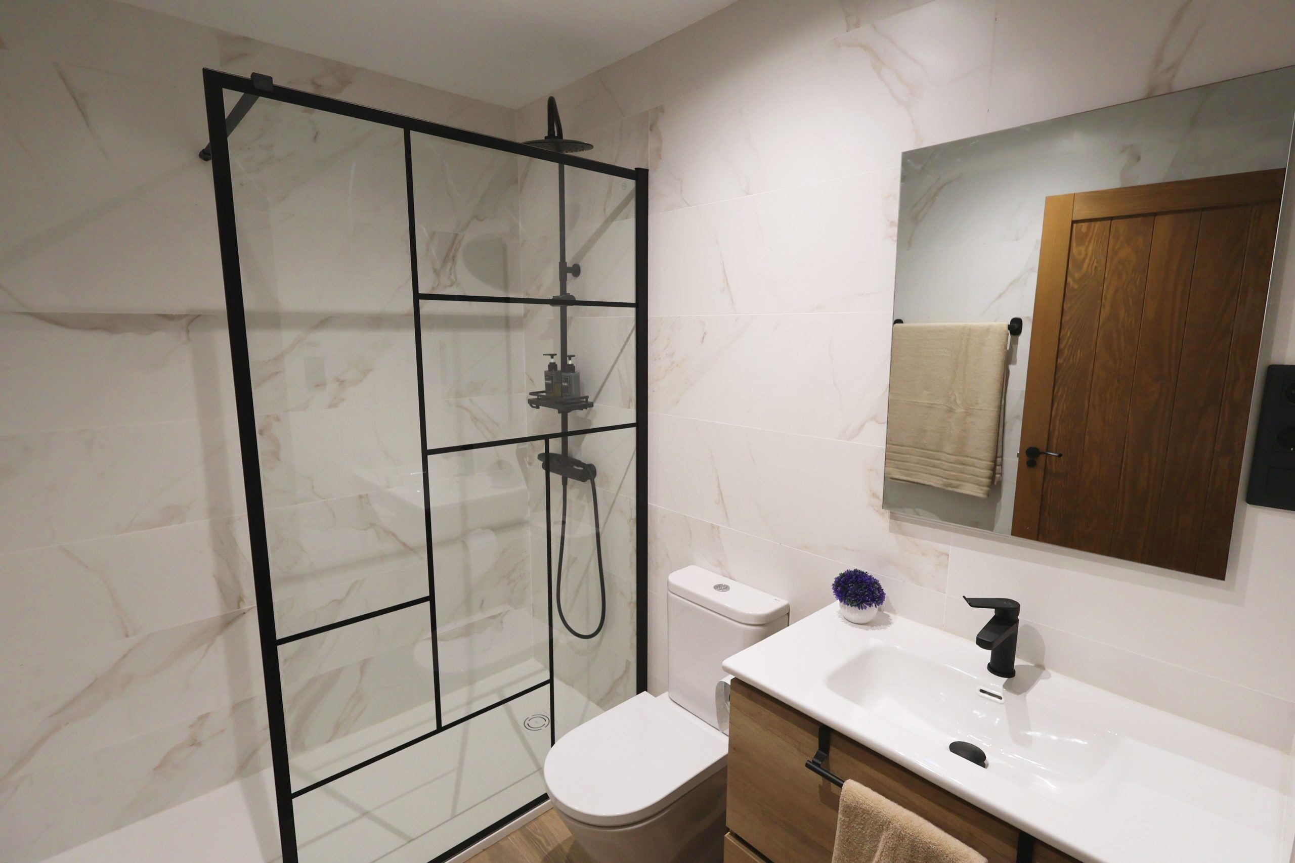Een moderne badkamer met een douchecabine van glas, een wit toilet, een wastafel met spiegel erboven en een houten kastje.