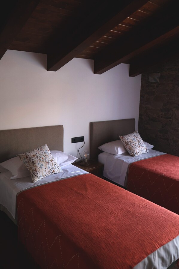 A hotel room with two beds, each with a red bedspread and white linens. The room has wooden beams on the ceiling and a stone wall.