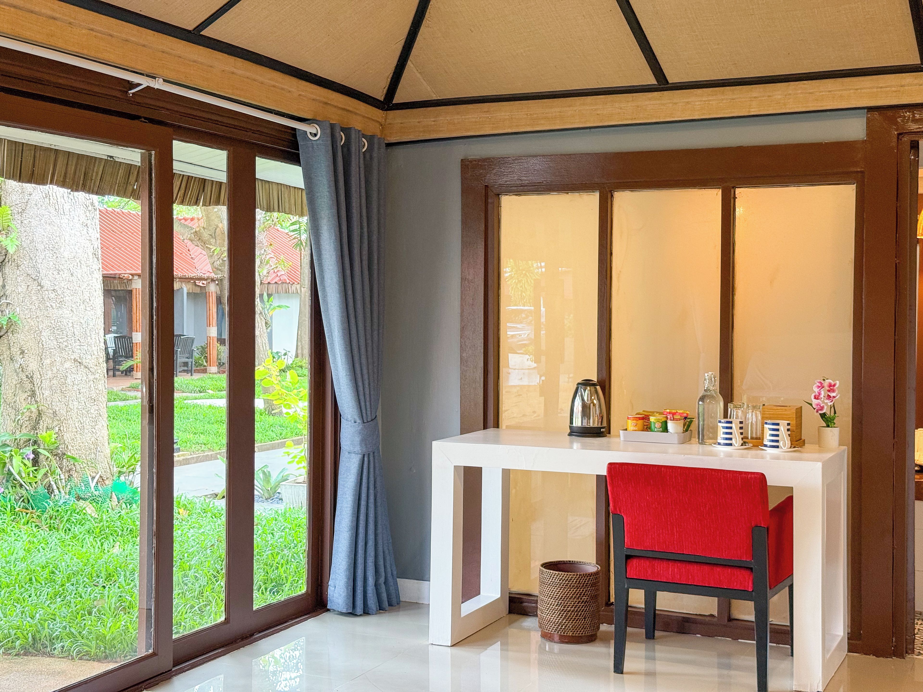 A room with a white table, a red chair, a window with curtains, and a view of greenery outside.