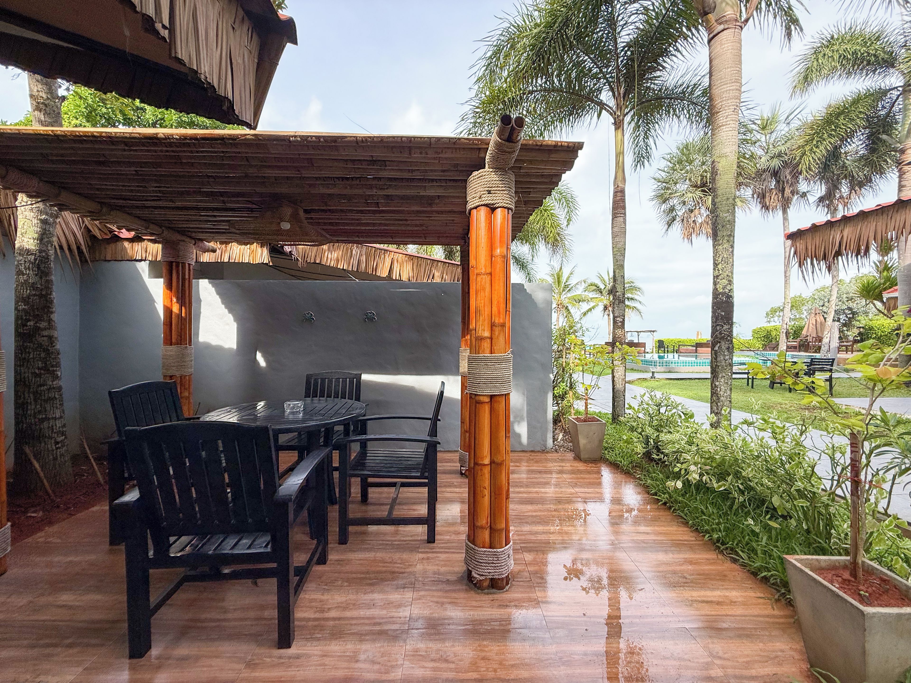 Un patio al aire libre con una mesa y sillas de madera, rodeado de macetas con plantas y palmeras.