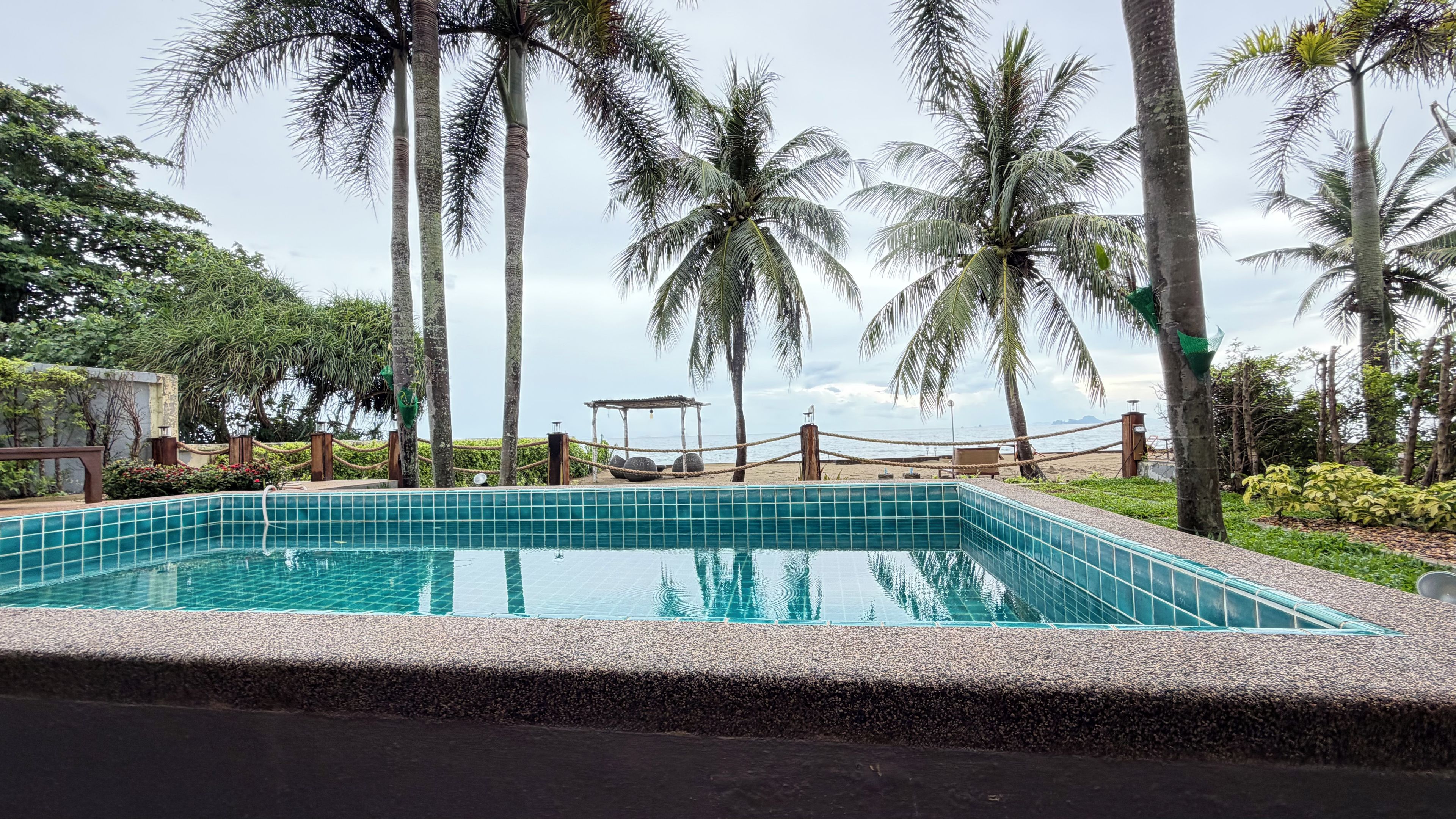 Una piscina rectangular con borde de azulejos, rodeada de piedras, palmeras y vista al mar.