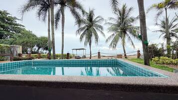 Una piscina rectangular con borde de azulejos, rodeada de piedras, palmeras y vista al mar.