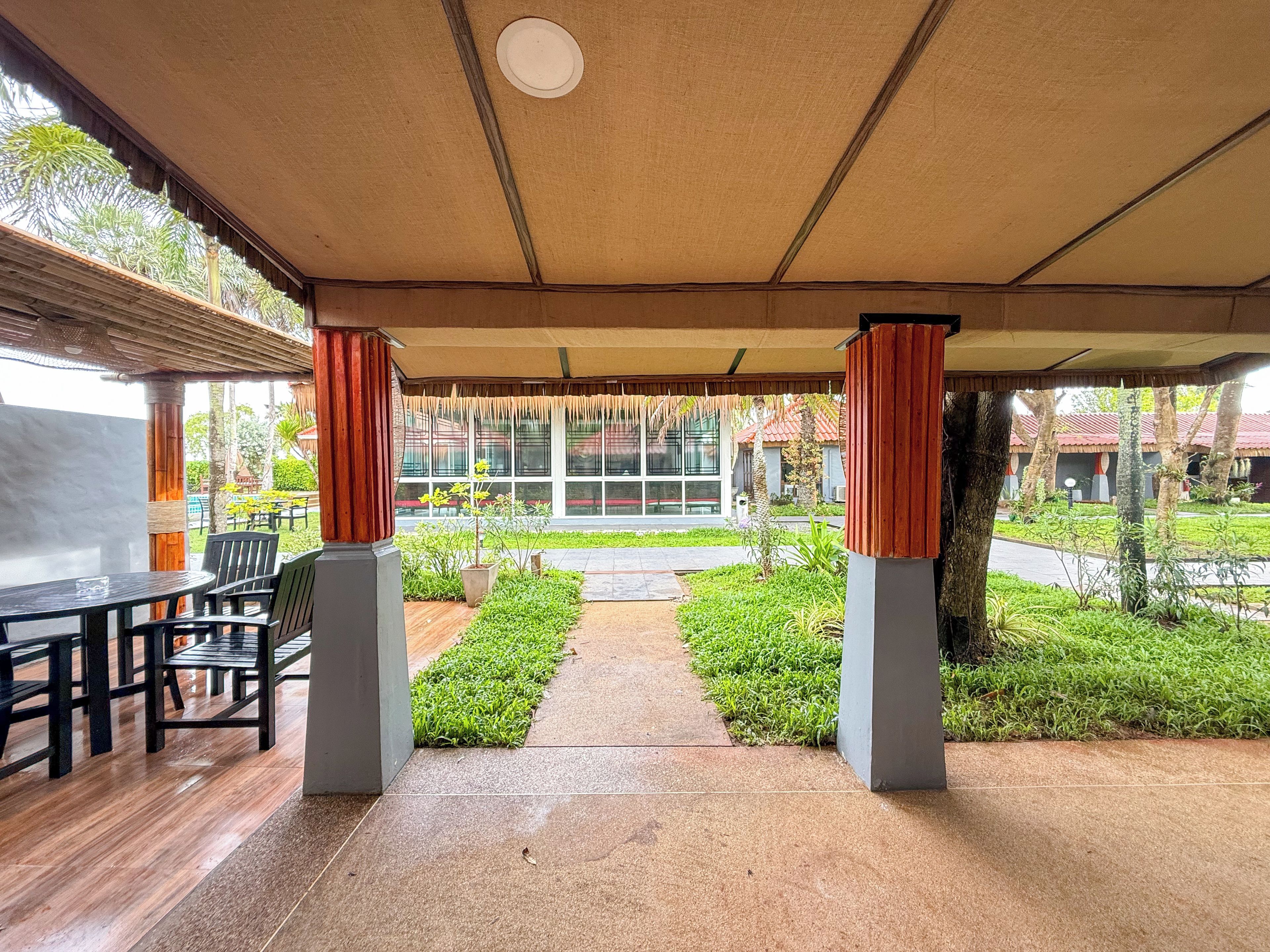 An outdoor covered area with a table and chairs, a pathway leading to a building, and greenery.