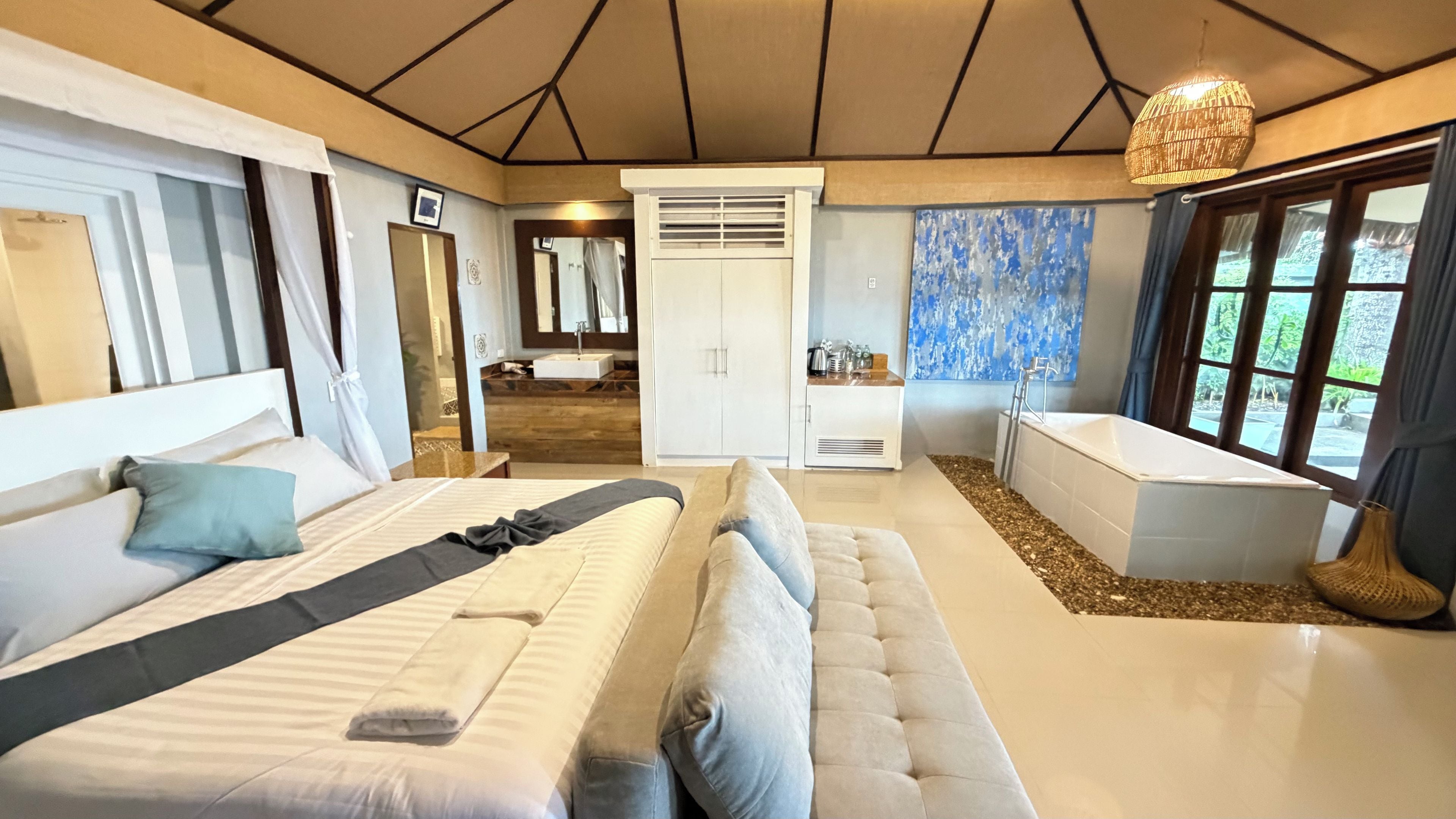 A modern bedroom with a large bed, a sitting area, a bathroom with a bathtub, and a view of the outdoors.