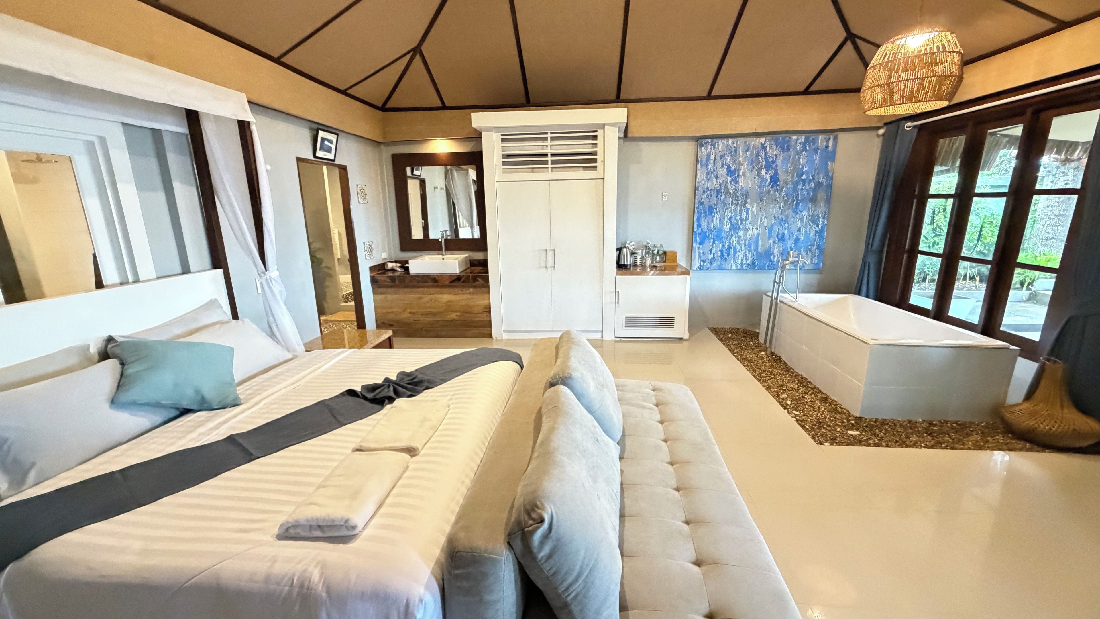 A modern bedroom with a large bed, a seating area, a bathroom with a bathtub, and a view of greenery outside.