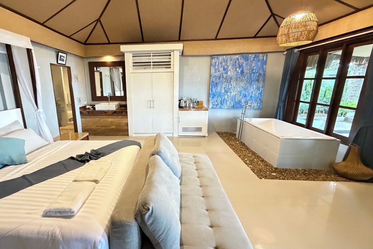 A modern bedroom with a large bed, a seating area, a bathroom with a bathtub, and a view of greenery outside.