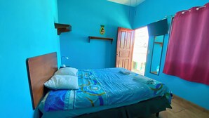 Ein Schlafzimmer mit Bett, Holz-Kopfteil, Nachttisch, Wandregal, Tür und Fenster mit Ausblick, sowie einem roten Vorhang.