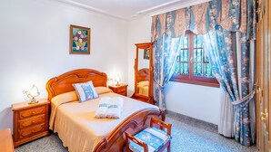 A hotel room with a wooden bed, a nightstand with a lamp, a mirror, and a window with floral curtains. - Cubo's Paraje Galiano Rural House / Private pool / BBQ / Free Wi-Fi (Guaro)
