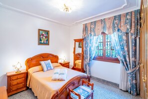 A hotel room with a wooden bed, a nightstand with a lamp, a mirror, and a window with floral curtains. - Cubo's Paraje Galiano Rural House / Private pool / BBQ / Free Wi-Fi (Guaro)