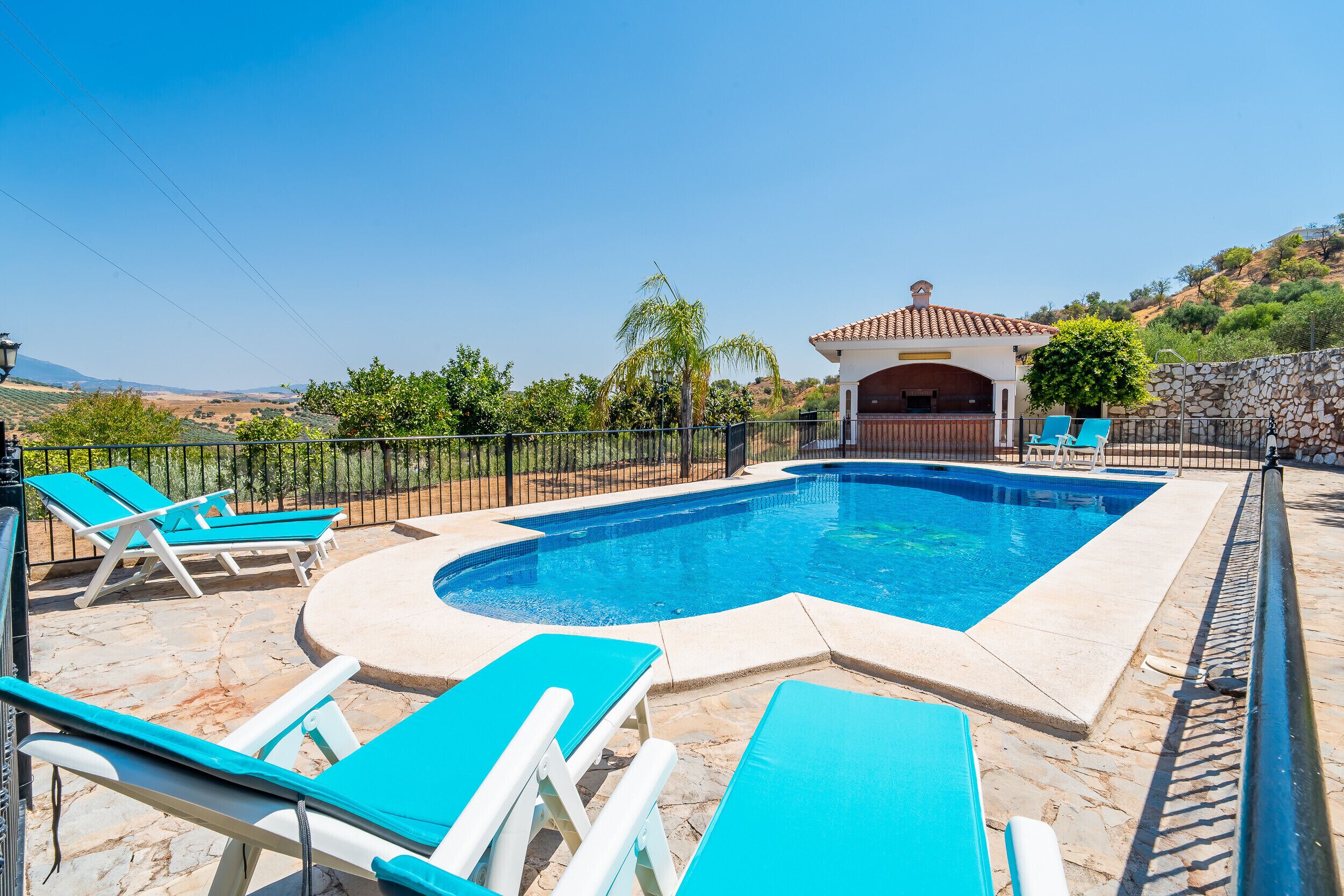 A swimming pool with blue water, surrounded by stone tiles and a stone wall. There are blue lounge chairs and a covered area with a tiled roof.