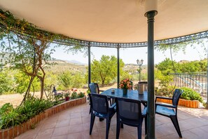 Ein überdachter Außenbereich mit Tisch und Stühlen, umgeben von Pflanzen und Blick auf die Landschaft.
