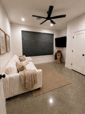 A modern living room with a sectional sofa, a ceiling fan, a large window with blinds, a flat-screen TV, and a vintage clock.