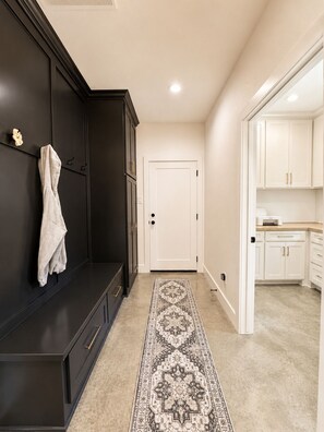 Un pasillo con un banco de color oscuro, una puerta blanca y una zona de cocina con gabinetes blancos y una encimera.
