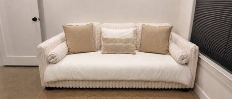 Ein weißes Sofa mit abgerollten Kanten und beigen Kissen, vor einer weißen Wand mit einem Fenster und horizontalen Jalousien.