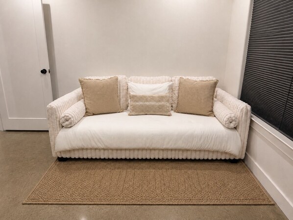 Un sofá blanco con bordes redondeados y cojines color beige, ubicado frente a una pared blanca con una ventana y persianas horizontales.
