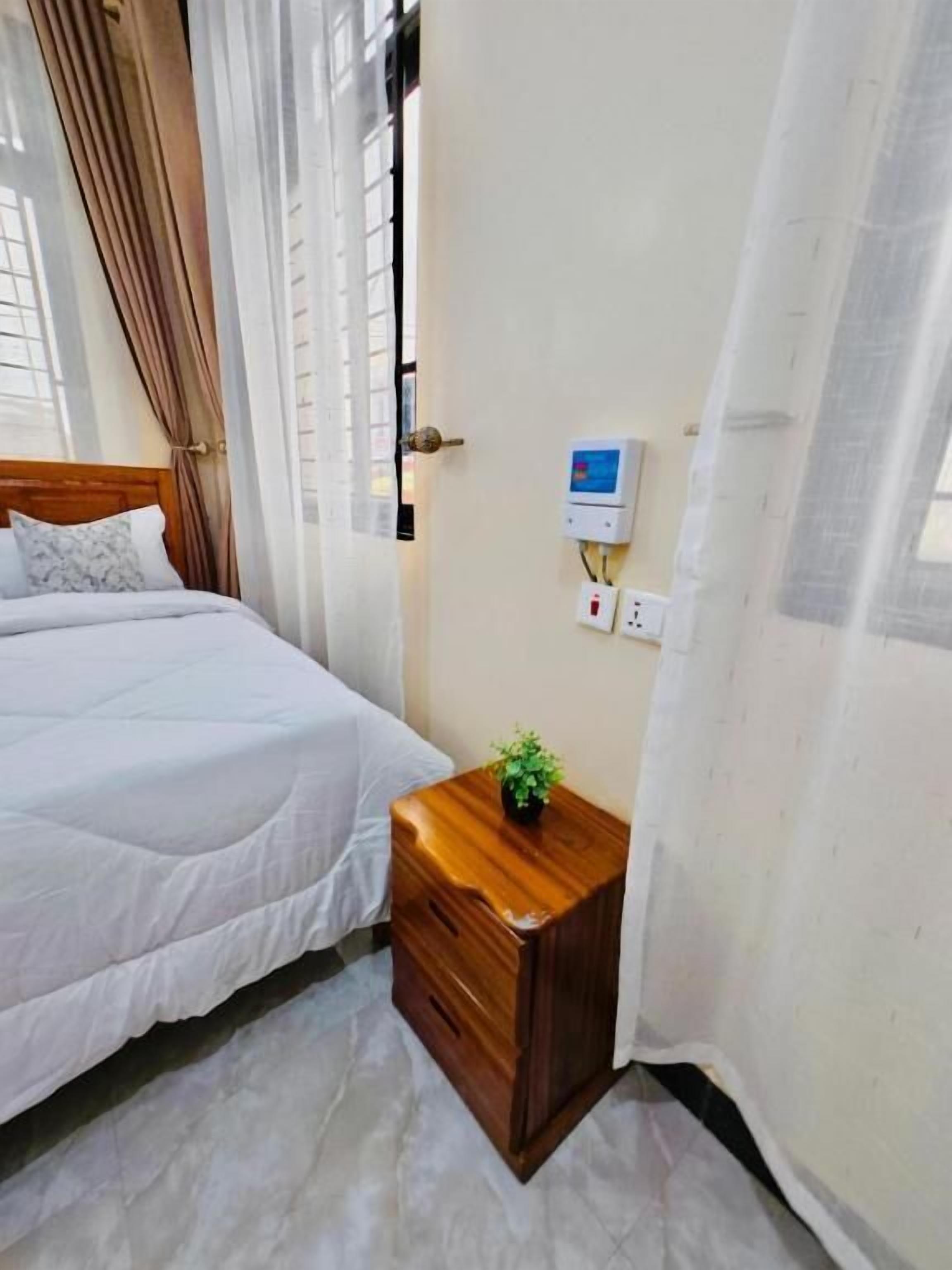 Un dormitorio con cama, mesita de noche, enchufe y ventana con cortinas.