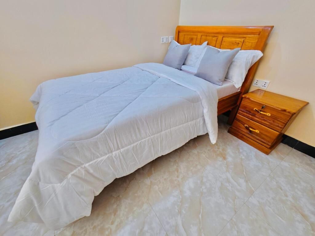 A neatly made bed with a wooden headboard, a bedside table with drawers, and a tiled floor.