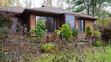 A wooden cabin with a porch, surrounded by deciduous trees and shrubs.