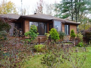 A wooden cabin with a porch, surrounded by deciduous trees and shrubs.