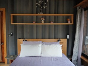 A bed with two pillows, a wooden headboard, a shelf above with decorative items, a small table, and a window with curtains. - Lovely 3-bedroom cottage with a 3-level treehouse (Woodbourne)