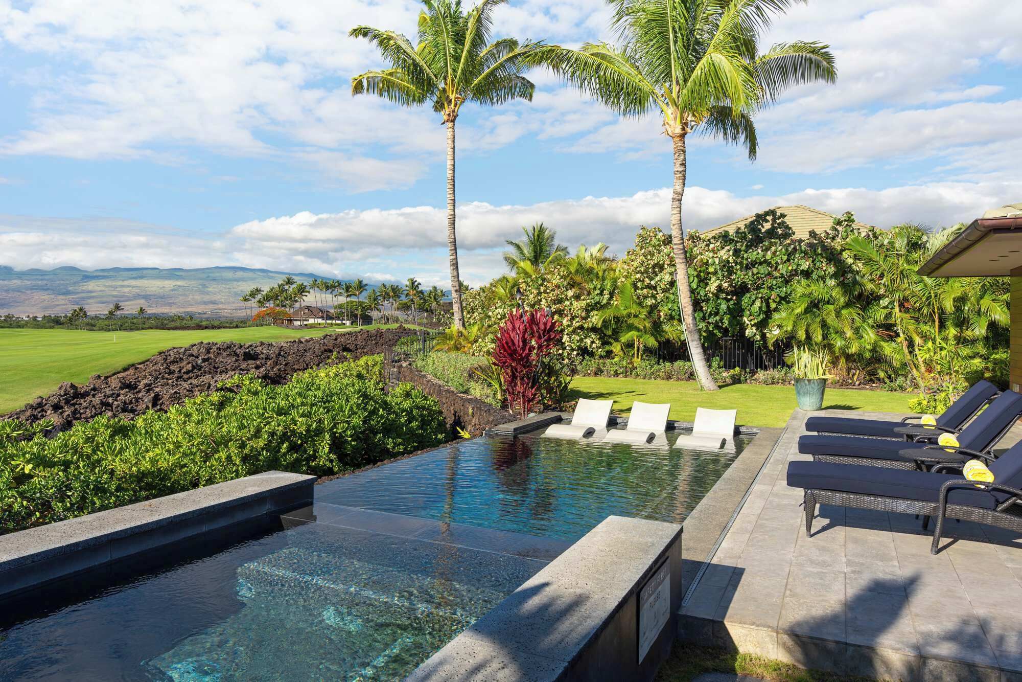 A swimming pool with clear water, surrounded by lounge chairs and palm trees, with a view of a golf course and mountains in the background.