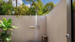 Un espacio de ducha exterior pequeño con un banco de madera, rodeado de plantas y una palmera.