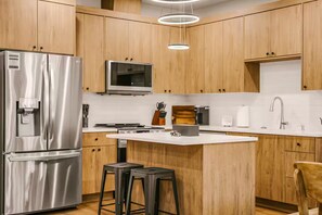 A modern kitchen with wooden cabinets, stainless steel appliances, a central island, bar stools, and a hanging light fixture. - Scandinavian 4BR - King Suite · EV Plug · Sleeps 10 (Gresham)