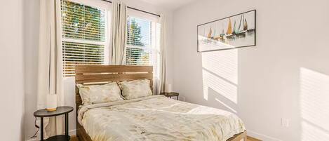 A bedroom with a bed, a nightstand, a bedside table, a window with blinds, and a painting on the wall.