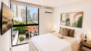Ein Schlafzimmer mit Bett, Nachttisch, Wand-Klimaanlage, Fenster mit Blick auf die Stadt und einem gerahmten abstrakten Gemälde.