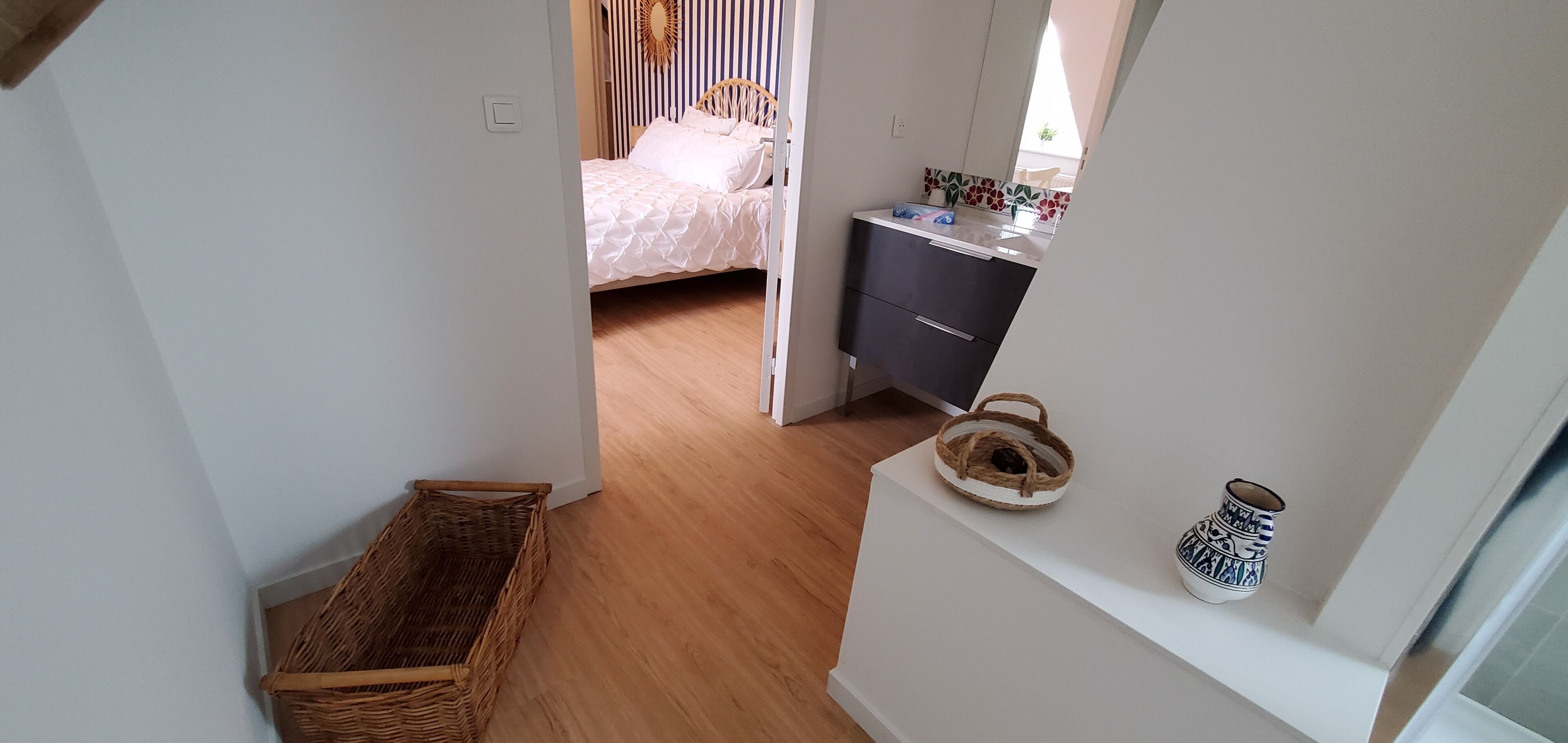 Um corredor estreito que dá para um quarto com cama, mesa de cabeceira e uma pequena janela.