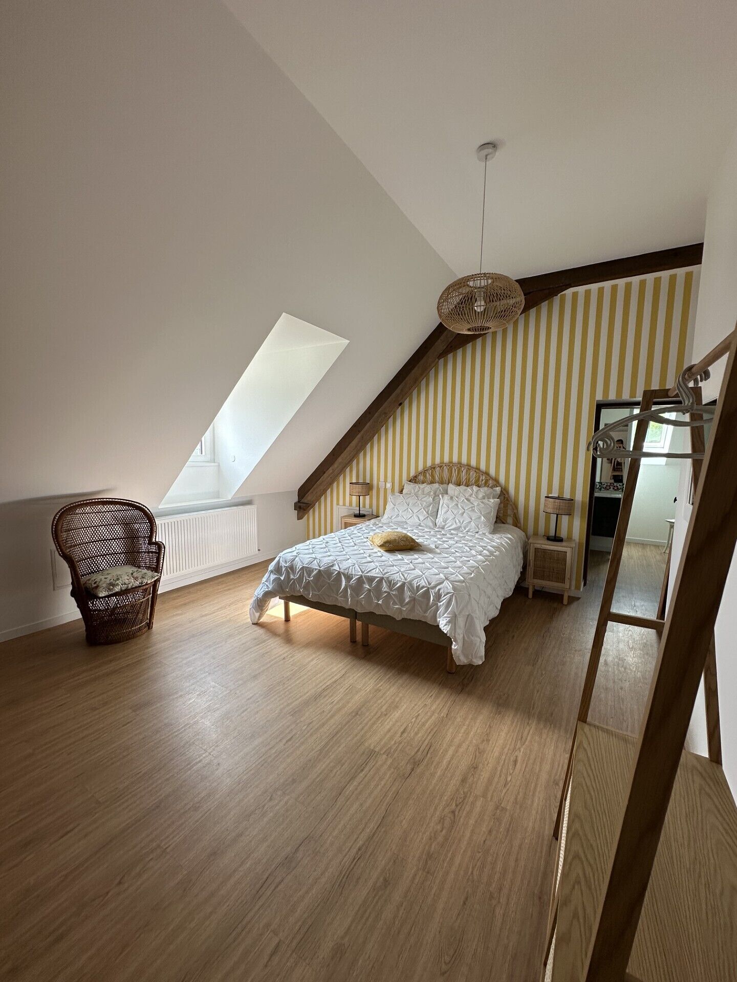 A bedroom with a sloped ceiling, a bed with a white duvet, a wicker chair, and a hanging light fixture.