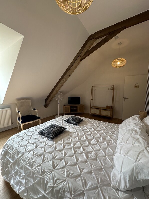 A bedroom with a sloped ceiling, a bed with a white quilted cover, a chair, a standing fan, a television, and a wooden wardrobe.