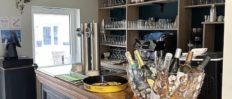 A bar area with a wooden counter, a large glass bowl filled with bottles, and shelves stocked with glasses and bottles.