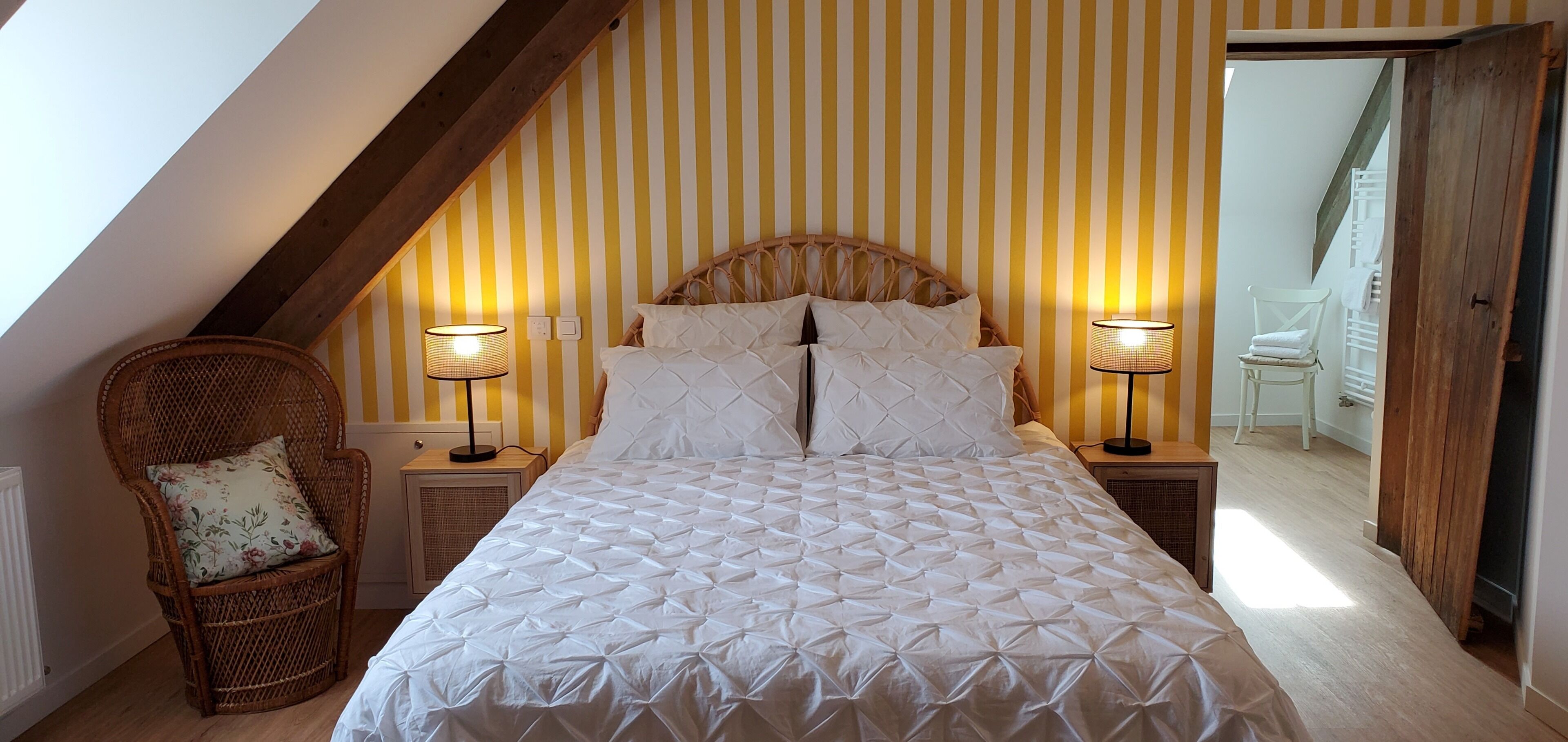 A bedroom with a sloped ceiling, a bed with a white quilted cover, two bedside lamps, a wicker chair with a floral cushion, and a wooden headboard.