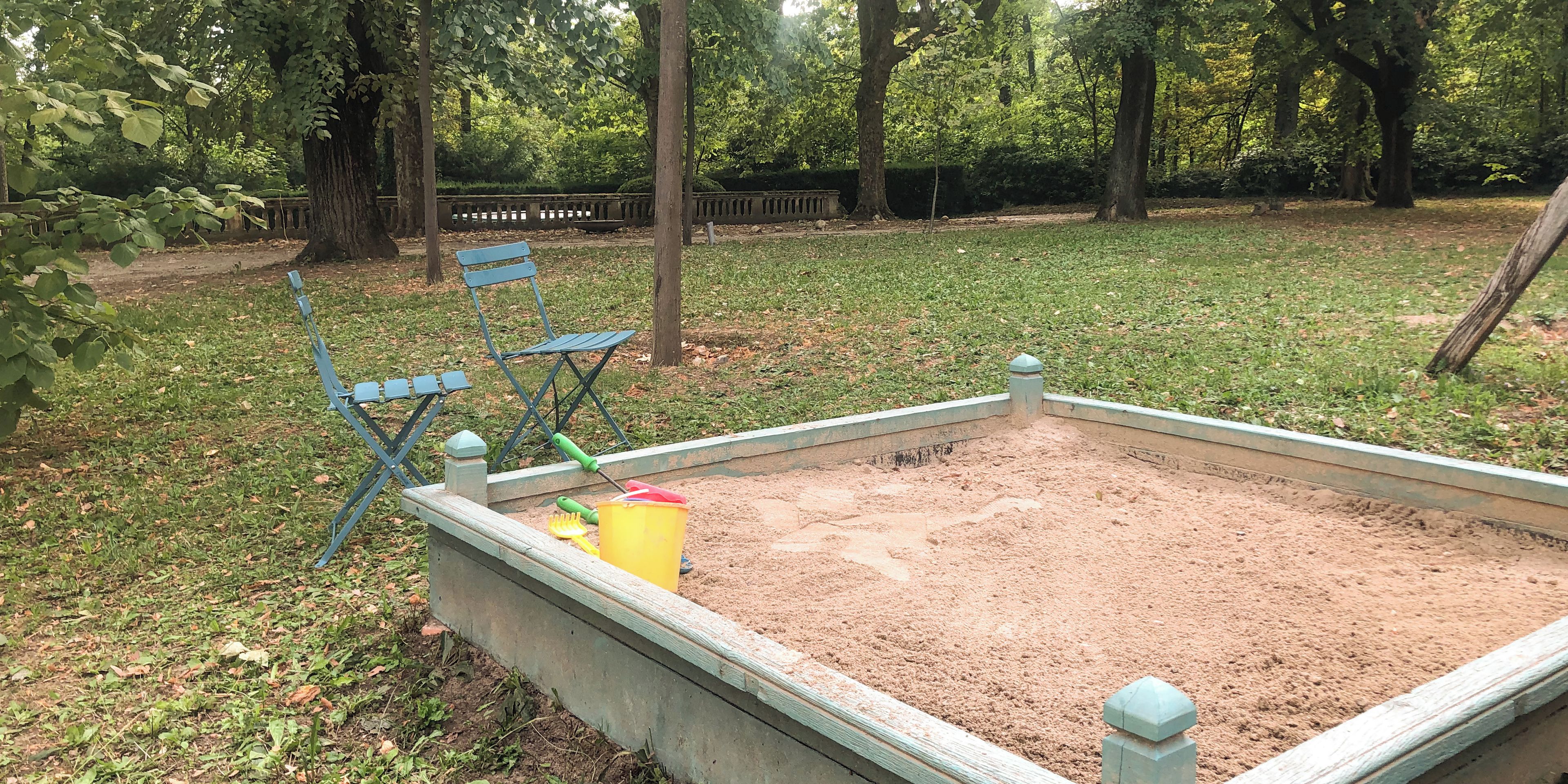 Ein Sandkasten mit Schaufel und Eimer, umgeben von Gras und Bäumen.