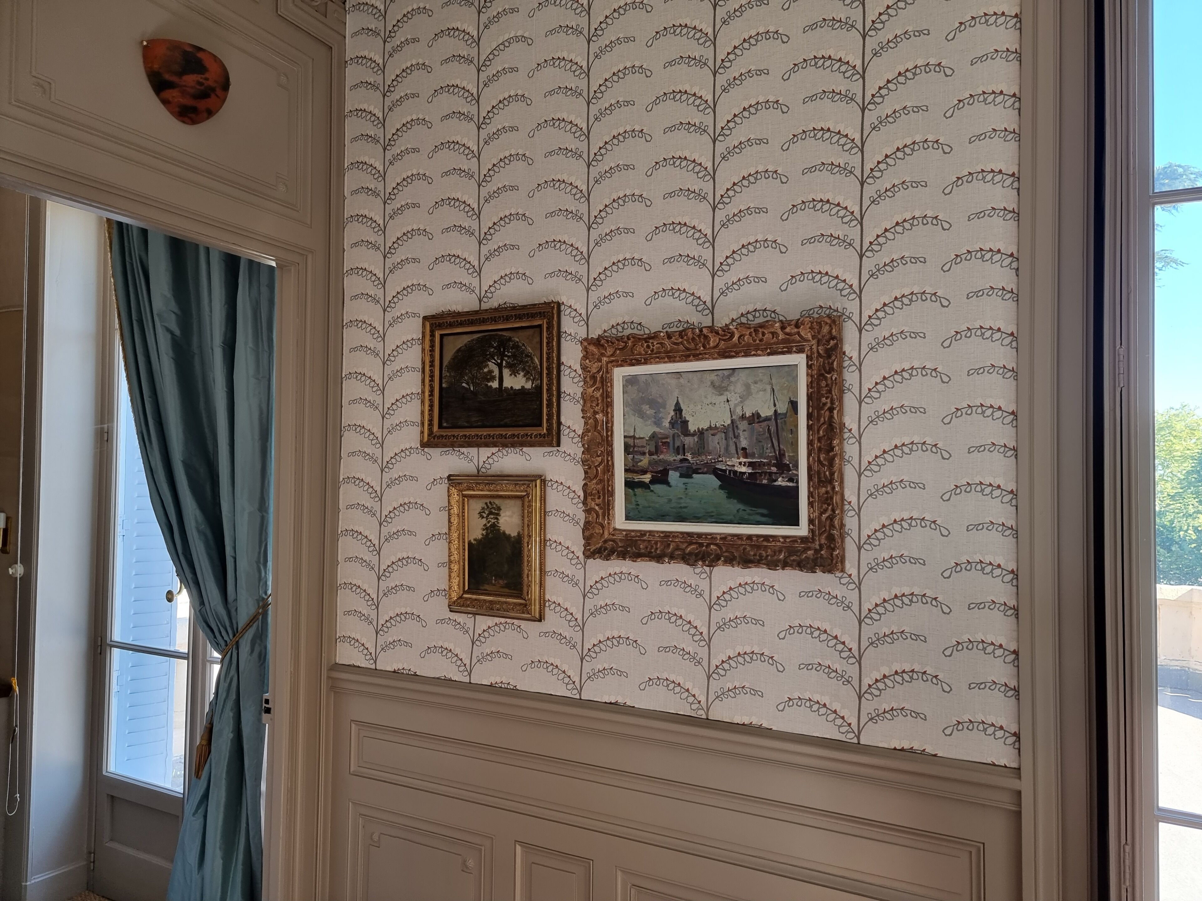 Een kamer met behang met patroon, een raam met blauwe gordijnen en drie ingelijste foto’s aan de muur.