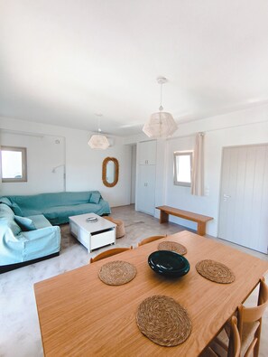 A modern living room with a sofa, a dining table with placemats, and a kitchen area with cabinets. - Persephone Sunset Villa (Paros)