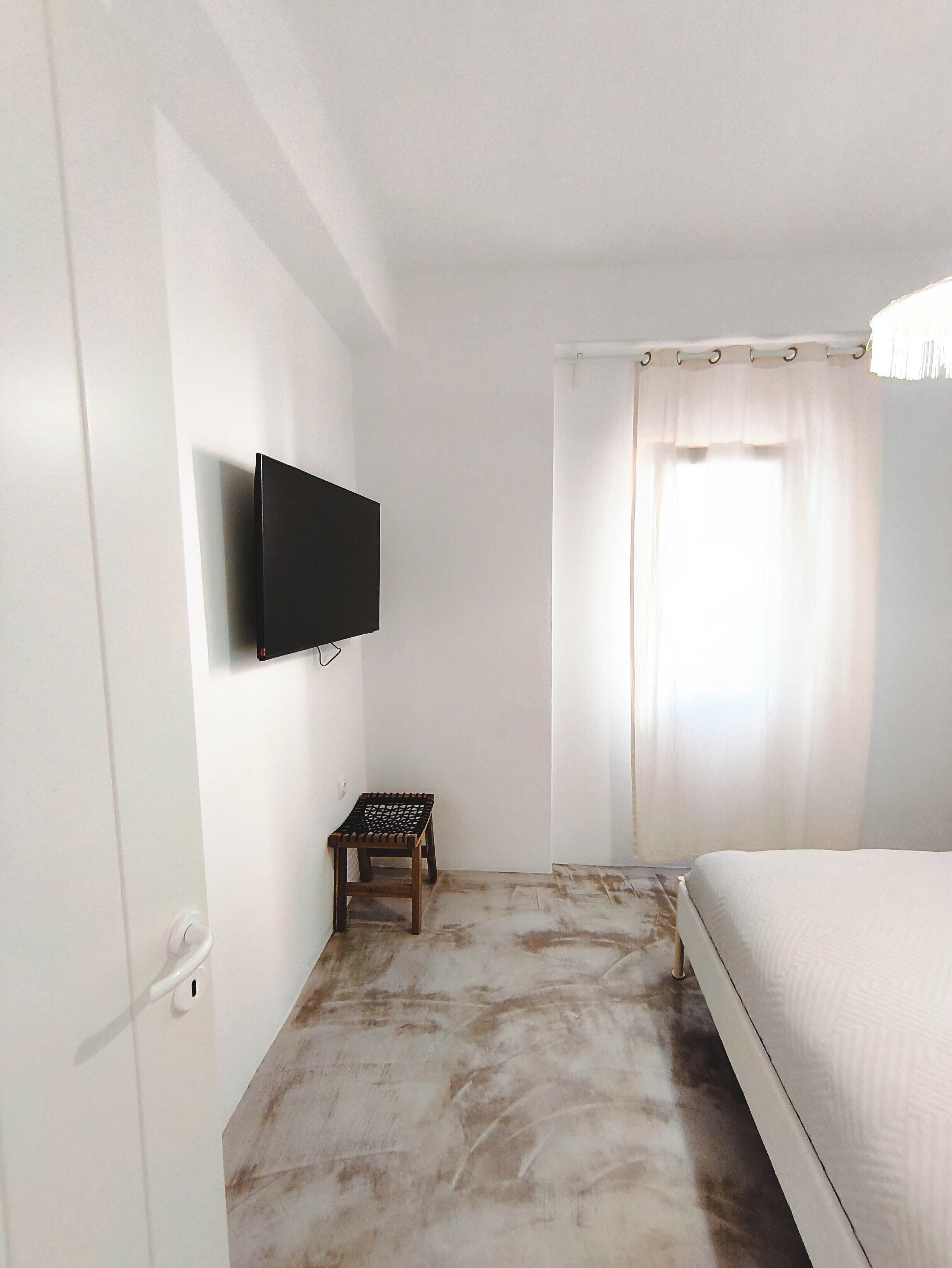 Une chambre avec un lit, un téléviseur fixé au mur, une petite table et une fenêtre avec des rideaux.
