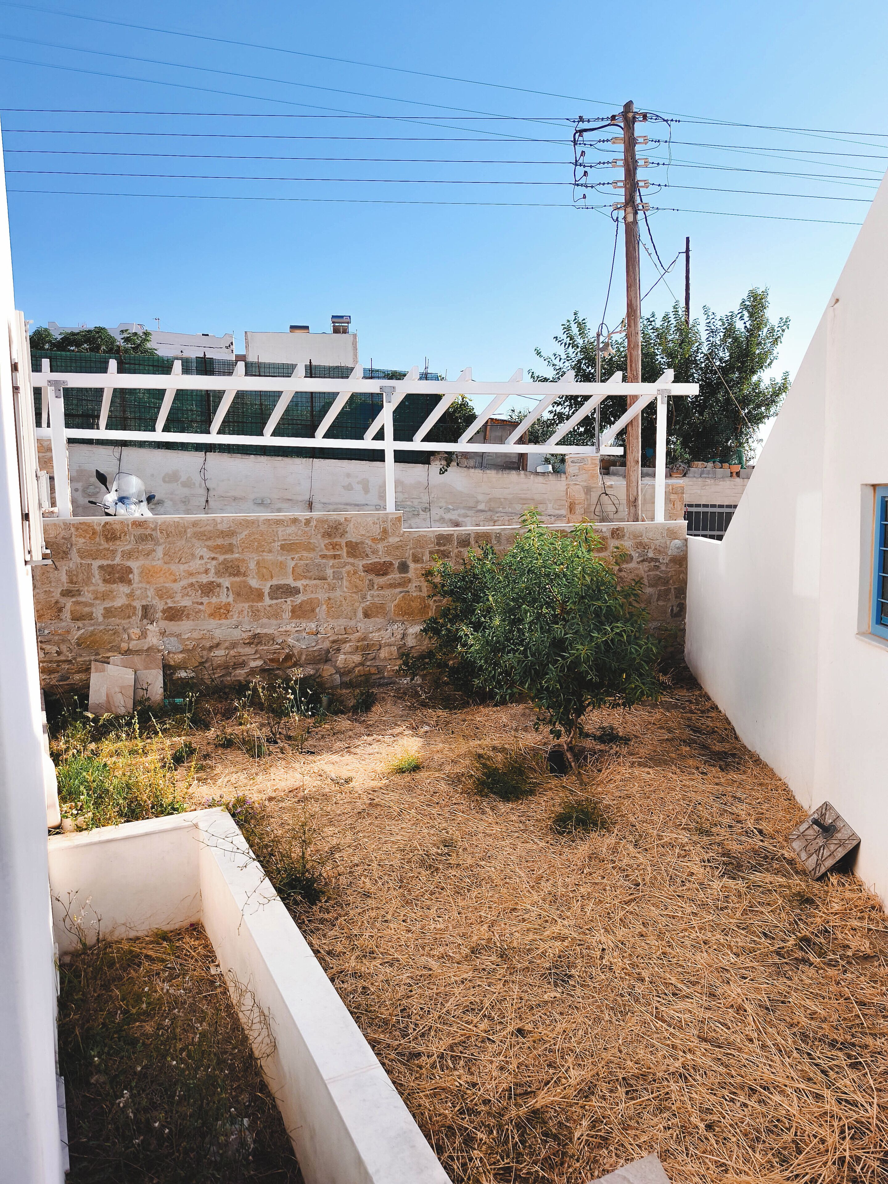 A courtyard with a white pergola, stone walls, and a small bush.