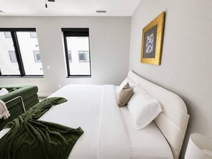 A bedroom with a large bed, a green chair, and a window. - Sleepover | Urban Studio + Gym/Sauna - Omaha (Omaha)