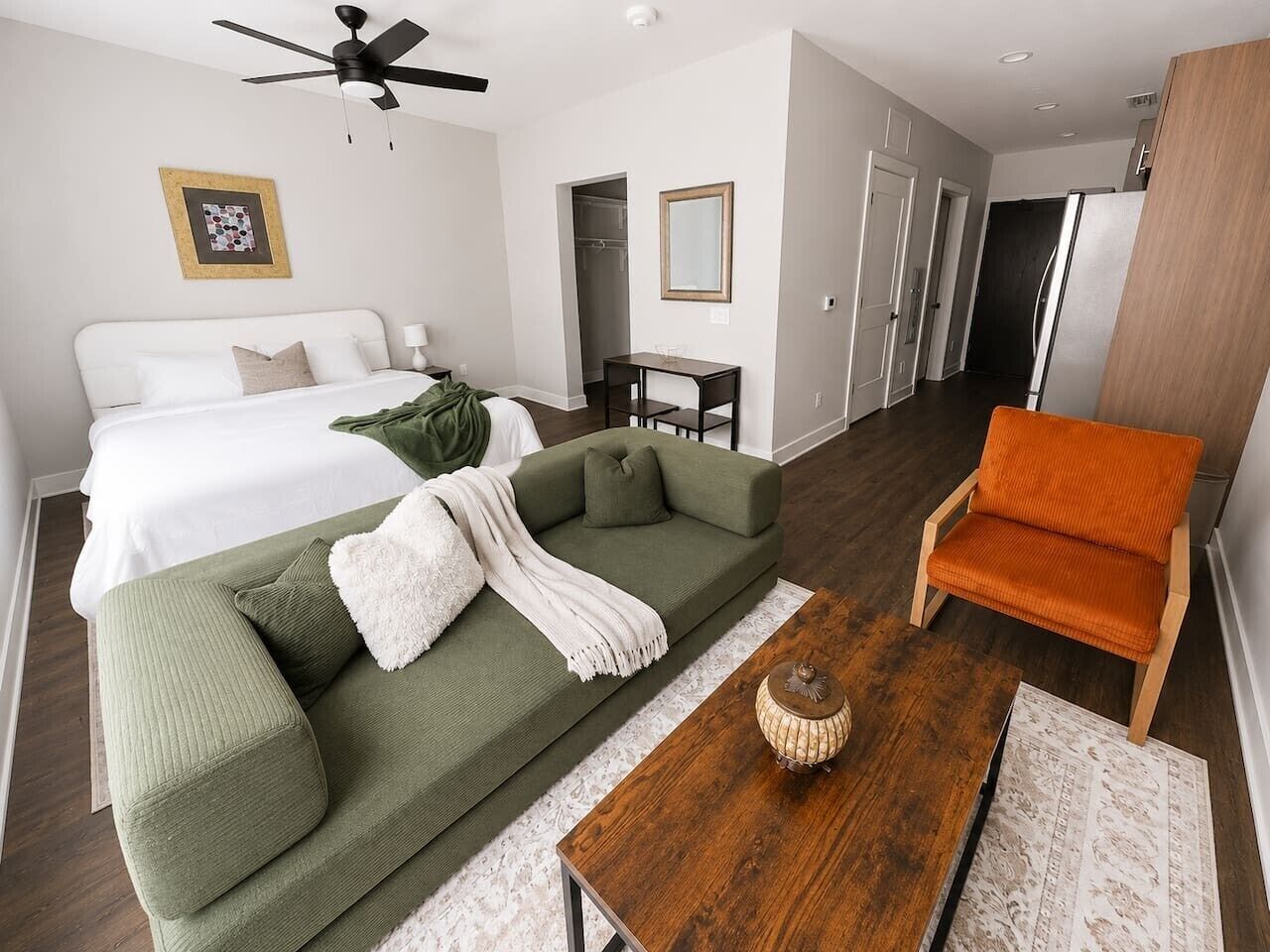 A modern hotel room with a bed, a green sofa, an orange chair, a wooden coffee table, and a ceiling fan.