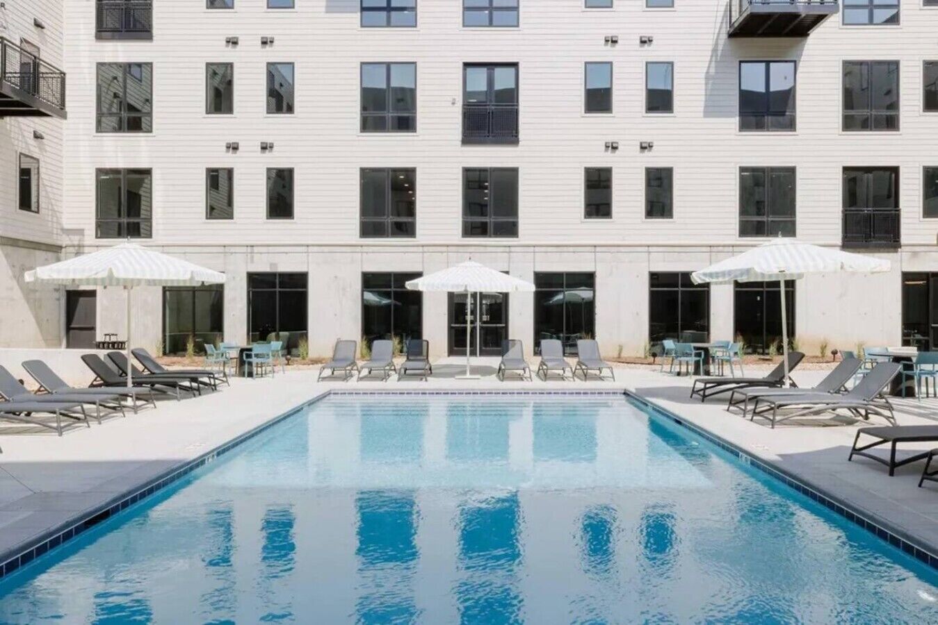 A modern building with a swimming pool, lounge chairs, and umbrellas.