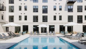 A swimming pool with lounge chairs and umbrellas, surrounded by a building with multiple windows and balconies. - Sleepover | Airy Studio + Gym/Sauna - Omaha (Omaha)
