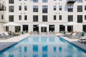A swimming pool with lounge chairs and umbrellas, surrounded by a building with multiple windows and balconies. - Sleepover | Airy Studio + Gym/Sauna - Omaha (Omaha)