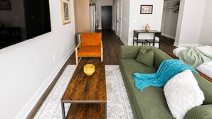 A modern living room with a flat-screen TV, a wooden coffee table, a green sofa, and a small table with a vase. - Sleepover | Airy Studio + Gym/Sauna - Omaha (Omaha)