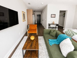 A modern living room with a flat-screen TV, a wooden coffee table, a green sofa, and a small table with a vase. - Sleepover | Airy Studio + Gym/Sauna - Omaha (Omaha)
