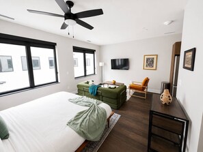 A modern bedroom with a large bed, a green sofa, a wooden side table, and a ceiling fan. - Sleepover | Airy Studio + Gym/Sauna - Omaha (Omaha)
