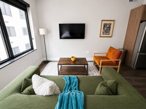 A living room with a green sofa, a wooden coffee table, a TV mounted on the wall, and a framed picture. - Sleepover | Airy Studio + Gym/Sauna - Omaha (Omaha)