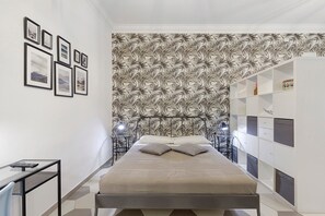 Ein Schlafzimmer mit einem Bett, Bildern in Rahmen an der Wand, Tapete mit Muster und einem weißen Kleiderschrank.