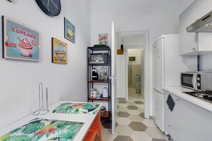 Eine Küche mit Mikrowelle, Gasherd und einem Regal mit Küchenutensilien. An der Wand hängt eine Uhr und es gibt eine Tür zu einem Badezimmer.